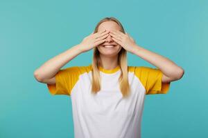 Pleased young attractive fair-haired lady with casual hairstyle covering her eyes with raised palms and smiling widely, standing over blue background in casual wear photo