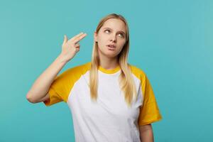 Discontent young pretty fair-haired woman with natural makeup raising hand with weapon gesture to her head and looking upwards with bored face, isolated over blue background photo