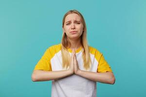 Wailful young pretty long haired blonde woman keeping her palms together and frowning sadly eyebrows while looking at camera, isolated over blue background photo