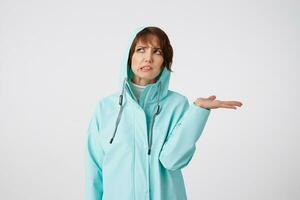 Photo of cute frowning short-haired curly woman in blue rain coat, hiding under the hood from rain and looking up at the left side, putting the palm under the rain standing over white background.