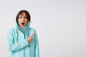 Shocked short-haired young woman in blue rain coat, with wide open mouth and outraged expression, wants to draw you attention to the copy space on the right side, stands over white background. photo