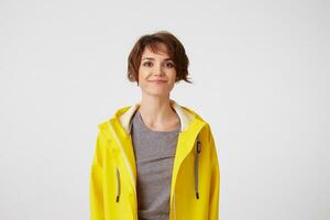 Photo of positive young nice woman in yellow rain coat, enjoy the life, looks at the camera with happy expressions, smiling over white wall.