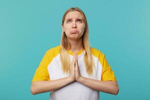 Portrait of young unhappy fair-haired female with long hair pursing her lips while looking sadly upwards, raising hands with praying gesture while posing over blue background photo