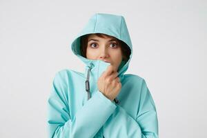 Close up of amazed young beautiful woman in blue rain coat, with a hood on his head, looks at the camera with surprise expressions, covers his face with a collar, stands over white wall. photo