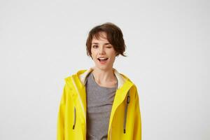 Photo of happy young nice woman in yellow rain coat, enjoy the life, looks at the camera with positive expressions and playful winks , broadly smiling over white wall.