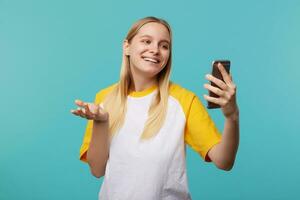 estudio foto de joven de cabeza blanca mujer con natural maquillaje sonriente alegremente a cámara mientras teniendo vídeo llamar, vestido en casual ropa mientras posando terminado azul antecedentes