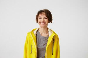 Young attractive girl in a yellow raincoat with a surprised expression on her face wants to draw your attention to the copy space on the left pointing with his finger, standing over white wall. photo