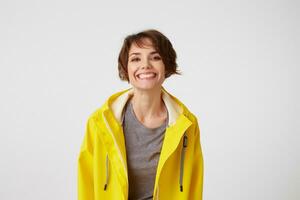 Photo of happy young nice woman in yellow rain coat, enjoy the life, looks at the camera with positive expressions, broadly smiling over white wall.