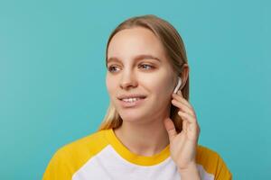 retrato de joven atractivo de ojos azules rubia largo peludo dama con auriculares mirando pensativamente aparte y sonriente suavemente, aislado terminado azul antecedentes foto