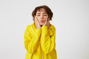 retrato de joven linda corto peludo niña usa en amarillo lluvia abrigo, enviar Beso con cerrado ojos y toques las mejillas, soportes terminado blanco pared. foto