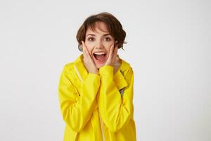 Portrait of young happy amazed cute short haired girl wears in yellow rain coat, with wide open mouth and eyes, touches cheeks, stands over white wall. photo