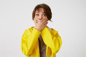 Portrait of scared short-haired curly woman in yellow rain coat, heard a terrible storry, covered mouth with palms, stands over white wall with wide open eyes with afraided expression. photo