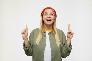 Cheerful young lovely white-headed lady with casual hairstyle keeping her forefingers raised and looking joyfully upwards, standing against blue background photo