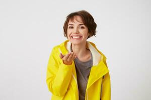 Portrait of young happy cute short haired girl wears in yellow rain coat, broadly smiles and pointing palms at the camera, like holding something small in the palm, stands over white wall. photo