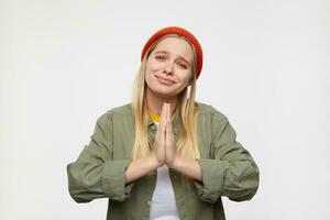 Portrait of young lovely long haired blonde woman keeping raised palms together while begging for something and looking drearly at camera, posing over blue background photo