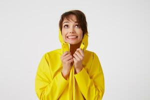 Portrait of young happy cute short haired girl wears in yellow rain coat, hiding under a rain hood, broadly smiles and looks up, stands over white wall. photo