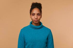 Close up of sad young African American girl wearing a blue sweater, with curly dark hair. Looking at the camera dropping his lips isolated over beige background. photo