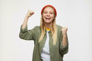 Studio photo of young long haired blonde lady raising happily fists and looking joyfully at camera, wearing red hat, olive shirt and white t-shirt while standing over blue background