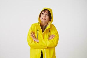 Portrait of young disgusted cute short haired girl wears in yellow rain coat, looks up with unhappy expressions, hiding under a rain hood, stands over white wall with crossed arms. photo