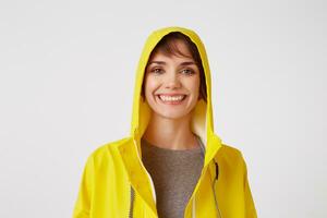 Close up of young attractive happy girl in a yellow raincoat, standing over white wall and broadly smiling. Enjoying the day. Positive emotion concept. photo