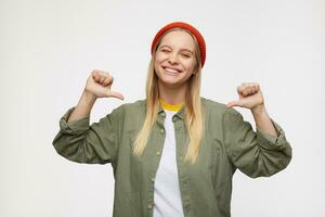 Indoor shot of young joyful white-headed female pointing cheerfully on herself with raised hands and giving wink at camera, isolated over blue background photo