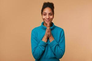 Portrait of young African American girl with curly dark hair wearing a blue sweater. Smiling, keeps palms together, pleads for mercy. Isolated over beige background. photo