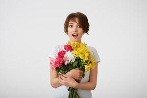 Portrait of happy amazed young cute short haired girl in white blank t-shirt, with wide open mouth and eyes, holding a bouquet of colorful flowers, surprised by such a gift, isolated over white wall. photo