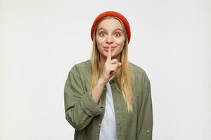 Positive young pretty long haired blonde female with casual hairstyle looking excitedly at camera and raising hand with hush gesture, isolated over blue background photo