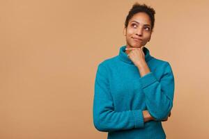 Photo of puzzled African American girl with curly dark hair wearing a blue sweater. Touches chin can't decide, doubts, look up isolated over beige background with copy space.