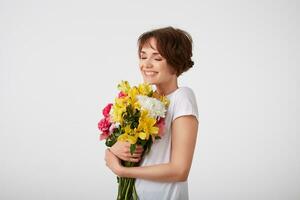 Portrait of happy cute young short haired lady in white blank t-shirt, holding a bouquet of colorful flowers, enjoying the smell with closed eyes, smiling broadly, standing over white background. photo