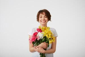 Happy cute young short haired girl in white blank t-shirt, holding a bouquet of colorful flowers with closed eyes, smiling broadly, standing over white background. photo