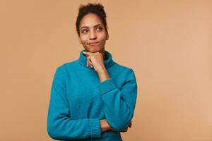 Photo of thinking young African American male with curly dark hair wearing a blue sweater. Touches chin, look up isolated over beige background with copyspace.