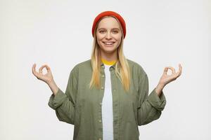 Portrait of attractive young white-headed female with natural makeup folding mudra gesture with raised hands and looking at camera with charming smile, isolated over blue background photo