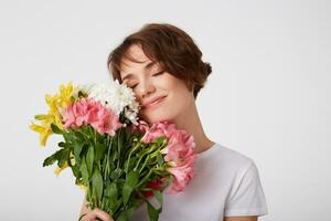 Photo of cute short haired lady in white blank t-shirt, holding a bouquet, covers face with flowers, enjoying the smell, standing over white background with closed eyes.