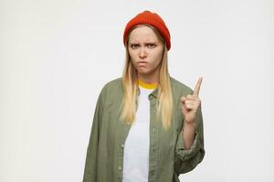 Severe young long haired blonde woman in red hat and casual wear keeping her forefinger raised while looking seriously at camera, standing over blue background photo