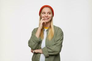 Indoor shot of young cheerful long haired blonde lady holding raised hand on her face and looking gladly aside, posing over blue background in casual clothes photo