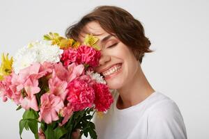 Portrait of nice short haired girl in white blank t-shirt, holding a bouquet, covers face with flowers, standing over white background with closed eyes. photo