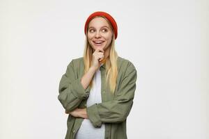 Cheerful young lovely white-headed female with natural makeup keeping raised hand on her chin while looking dreamily aside with wide smile, posing over blue background photo