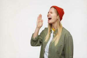 Excited young pretty long haired blonde lady keeping her eyes closed while screaming and raising hand to her mouth, wearing casual clothes while posing over blue background photo