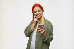 Glad young pretty long haired blonde woman with natural makeup holding mobile phone in raised hand and smiling while checking social networks, isolated over blue background photo