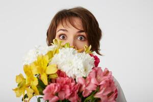 Portrait of nice short haired girl in white blank t-shirt, holding a bouquet, peeping from behind flowers, standing over white background with wide open eyes. photo