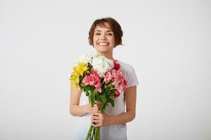 Portrait of smiling nice short haired lady in white blank t-shirt, holding a bouquet of colorful flowers, very glad and smiling over white wall. photo