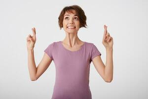 Young attractive young short-haired lady in blank t-shirt, looks up, bites lip, hopes for luck with crossed fingers. Stands over white background. photo