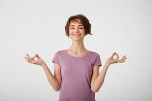 Concentrated young attractive young short-haired lady in blank t-shirt, keeps hands in okay gesture, meditates indoor, has eyes closed, tries to relax, isolated on white wall. photo