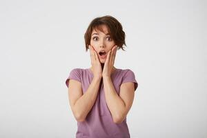 Portrait of wondered young attractive woman with a surprised expression on her face, standing over white wall with wide open moth and eyed. Positive emotion concept. photo