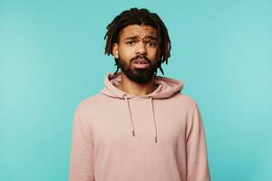 Bewildered young brown-eyed brunette bearded male frowning his eyebrows while looking confusedly ar camera, isolated over blue background in sporty wear photo