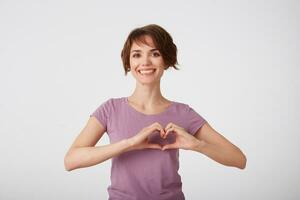 Beautiful cheerful short-haired lady in blank t-shirt, shows heart shape gesture, wears casual t shirt, being glad, stands over white background. Love symbol, romantic mood. photo