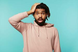 Indoor shot of young dark haired bearded male dressed in pink hoodie holding raised hand on his head while looking dazedly at camera, isolated over blue background photo