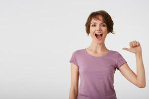 Self assured cheerful short-haired lady in blank t-shirt feels proud of her deeds, points at herself feels surge of pride, raises head, broadly smiles, wears casual clothes, isolated over white wall. photo