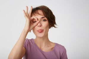 Close up of young attractive funny short-haired lady in blank t-shirt, standing over white wall, looking through okay gesture and shows tongue. Positive emotion concept. photo